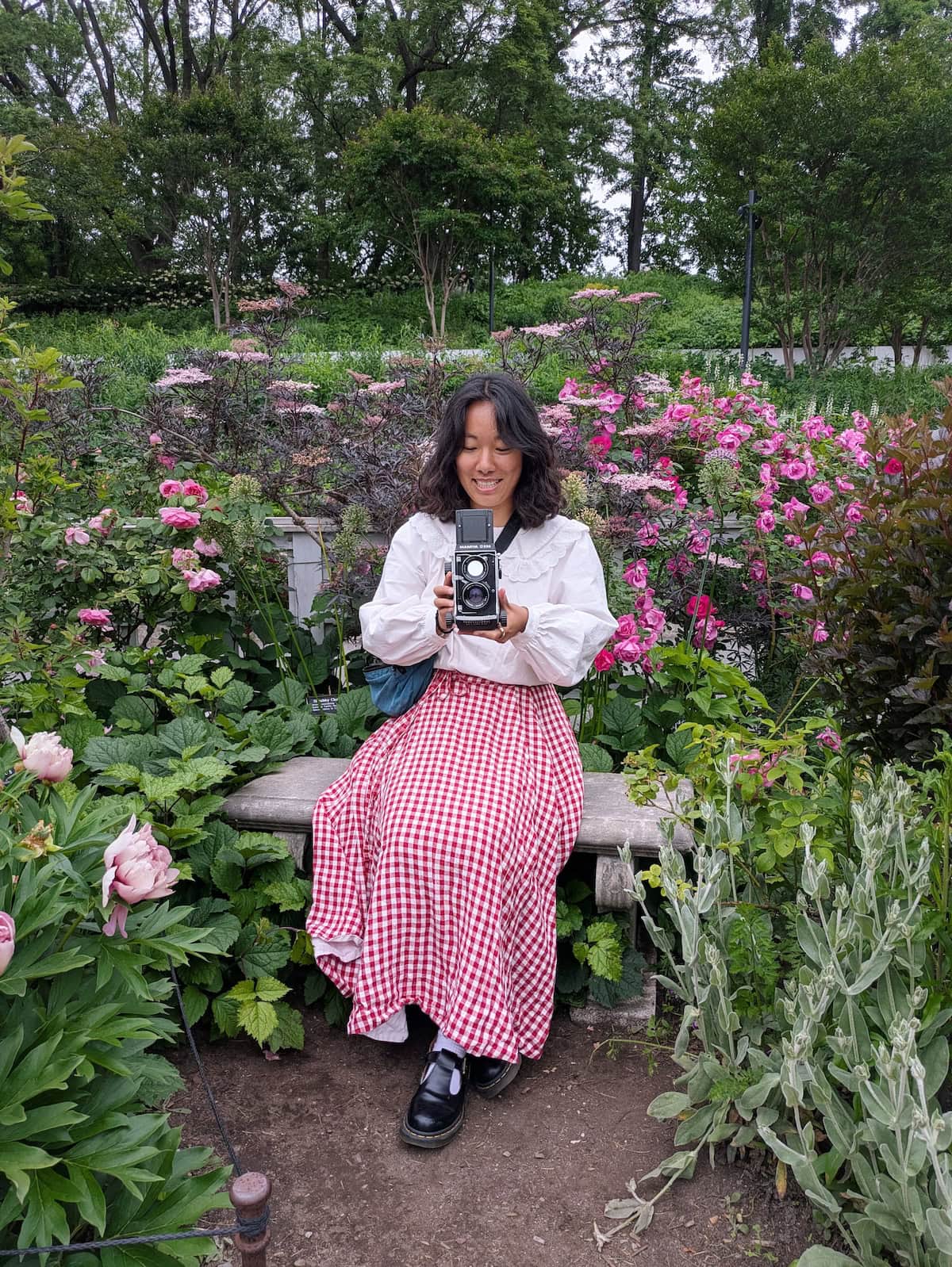 Me wearing the red gingham wrap skirt from Son de Flor sitting on a bench in a garden and holding a vintage camera